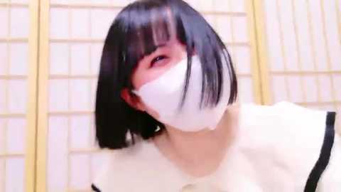 Media: Video of an East Asian woman with straight, black hair in a bob cut, wearing a white surgical mask, white top, and black shoulder straps. The background features a light-colored, grid-patterned screen.