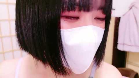 Media: Video of an Asian woman with straight black bob hair, wearing a white surgical mask, in a bathroom with a white tiled wall and a hanging towel.