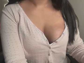 Media: Video of a woman with light skin and long black hair, wearing a cream ribbed cardigan with a deep V-neck, exposing ample cleavage. The background is blurred, focusing on her upper body.