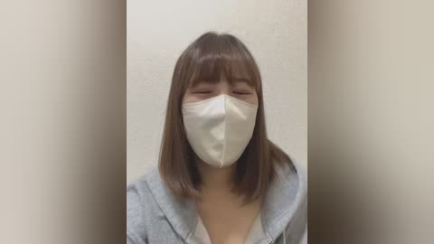 Media: A video of an East Asian woman with straight brown hair, wearing a white surgical mask, a light gray hoodie, and standing against a plain beige wall.