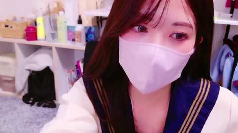 Media: Video of an Asian woman with long dark hair, wearing a white surgical mask, navy blue sailor collar top, and seated in a cluttered, brightly-lit room with shelves of colorful toiletries and a black bag on the floor.