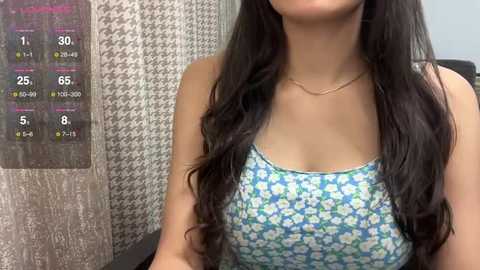 Media: Video of a young woman with long, wavy black hair, wearing a sleeveless blue floral dress, standing in front of a houndstooth-patterned wall with a height chart on the left.