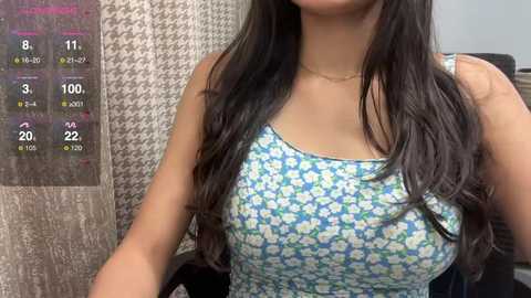 Media: Video of a woman with long, dark hair, wearing a blue floral sundress, standing in front of a houndstooth-patterned wall.