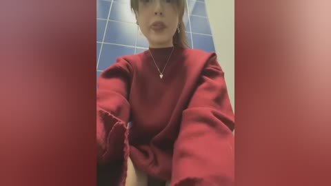 Media: Video of a young woman with fair skin, wearing a red sweater and a necklace, standing in front of blue-tiled walls. Her hair is dark and tied back.