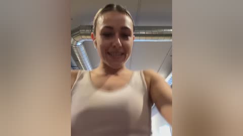 Media: A video of a smiling woman with light skin and blonde hair, wearing a tight, white tank top, standing in a modern room with exposed ductwork and gray walls.