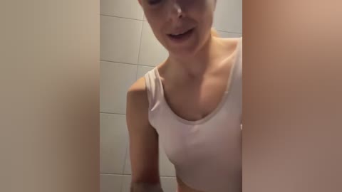 Media: Video of a smiling woman with light skin, wearing a white tank top, standing in a tiled bathroom with beige walls. Her face is partially obscured by the foreground, creating a sense of intimacy.