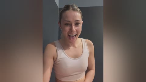 Media: Video of a fit, smiling woman with light skin and brown hair in a ponytail, wearing a white sports bra, standing in front of a blurred, dark grey wall.