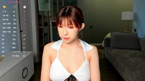 Media: Video of a young East Asian woman with short dark hair, wearing a white halter top, sitting indoors with a gray couch in the background.