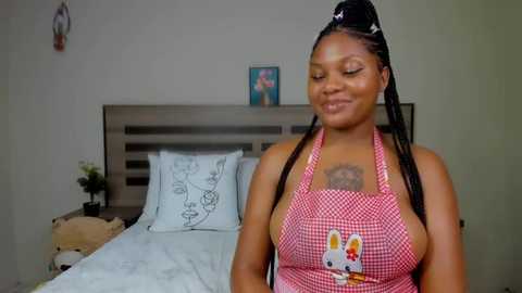 Media: Video of a smiling Black woman with braided hair, wearing a red apron with a bunny design, standing in a simple, modern bedroom with a wooden headboard and framed picture on the wall.