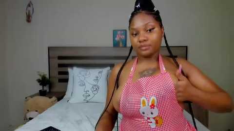 Media: Video of a Black woman with braids, wearing a red and white checkered bunny apron, standing beside a neatly made bed in a modern bedroom.