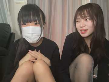 Media: Video of two young Asian women with long black hair, wearing black outfits, masks, and seated close together on a sofa, smiling. Background includes a dark chair and light-colored curtains.