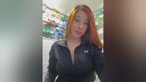 Media: Video of a young Asian woman with long orange hair, wearing a black Under Armour zip-up hoodie, standing in a brightly lit, modern kitchen with colorful food products on shelves in the background.