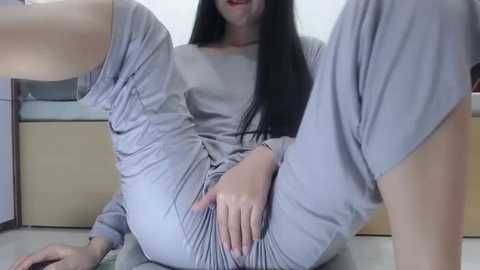 Media: Video of an Asian woman with long black hair, light skin, and a slim physique, wearing loose gray pajamas, seated on a tiled floor with legs spread, hand on her crotch.