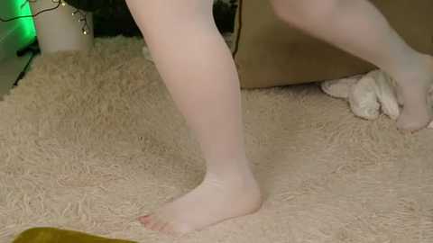 Video of a person wearing sheer white stockings, standing on a beige carpet, with a plush toy and a green light in the background.