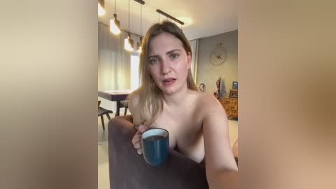 Media: Video of a topless blonde woman with medium breasts, holding a blue mug, seated in a modern living room with pendant lights, wooden furniture, and a clock on the wall.