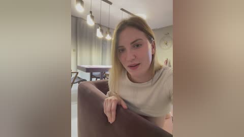 Media: Video of a blonde Caucasian woman with fair skin, wearing a beige top, leaning on a dark maroon chair in a modern, well-lit room with hanging lights, a dining table, and a painting on the wall.