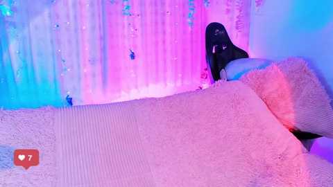 Media: A video of a person lying on a soft, fluffy white rug in a room with pastel pink and blue lighting. The person's body is partially covered, and a black object is visible in the background.