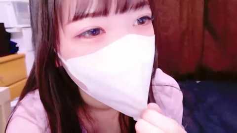 Media: Video of an East Asian woman with long dark hair and bangs, wearing a white surgical mask, in a room with wooden walls and a white cabinet.