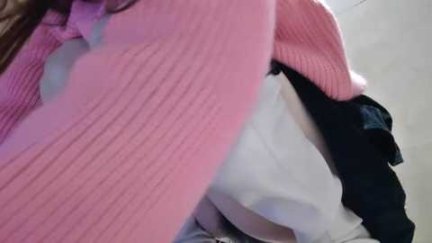 Media: Video of a person wearing a pink knitted sweater, partially covered by a white garment, with a black ruffled skirt visible. Background is blurred and neutral-colored.