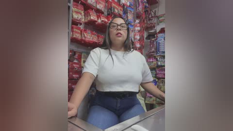 Media: Video of a plus-sized Latina woman with long black hair, glasses, and a white crop top, seated behind a checkout counter. The background features colorful packaged goods.