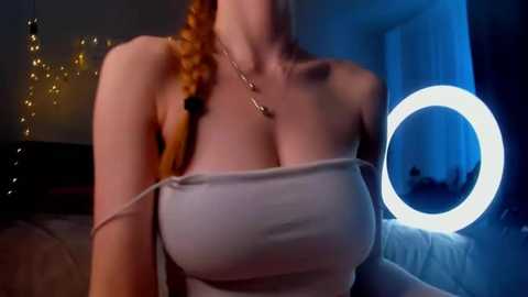 Video of a light-skinned woman with auburn hair, wearing a strapless white top, in a dimly lit room with a round light and fairy lights.