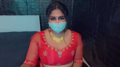 Media: A video of a South Asian woman in a red, embroidered saree with a gold necklace, wearing a blue surgical mask and sitting against a dark brick wall.