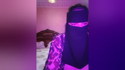 Media: A video shows a person wearing a black burqa with a pink floral pattern, standing in a dimly lit room with purple lighting. The background reveals a bed with brown and white sheets.