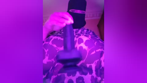 Media: A video of a person with light skin and medium-length blonde hair, wearing a black mask, holding a large, black dildo. The background is a bed with a leopard print blanket, and the lighting is purple.