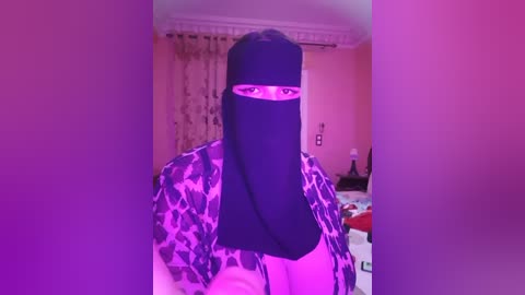 Media: A video of a person in a black niqab with a white patterned robe, standing in a room with pink lighting, visible curtains, and a bed in the background.