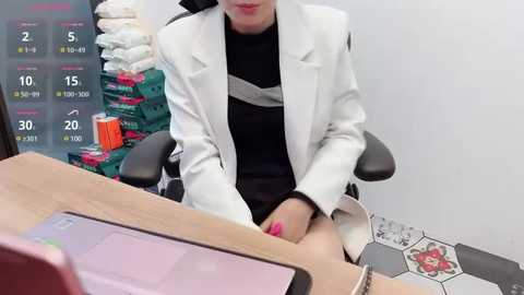 Media: Video of a stylish woman in a white blazer over a black turtleneck, sitting at a wooden desk with a smartphone and colorful stationery in the background.