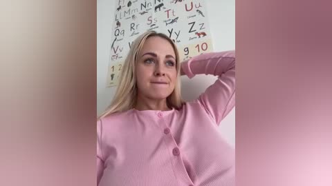 Media: Video of a blonde woman in a pink ribbed cardigan, posing against a wall with a periodic table poster, smiling slightly, with soft lighting.