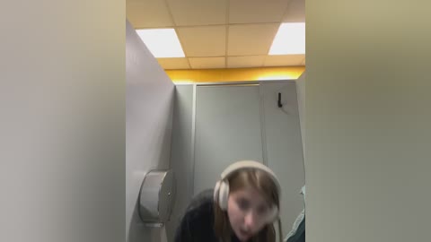 Media: A video of a dimly lit, narrow bathroom with a woman wearing a beige hooded jacket and headphones, blurred, standing next to a gray paper towel dispenser and yellow ceiling.