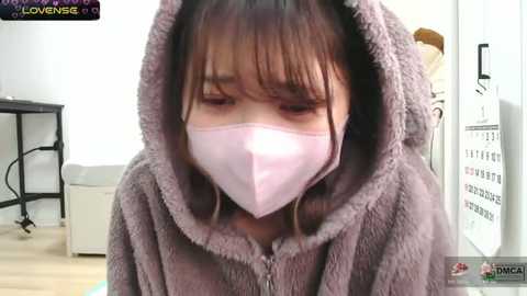 A video of an Asian woman with long, straight brown hair, wearing a gray, fluffy hooded robe and a white face mask, indoors with a black shelf and white wall in the background.