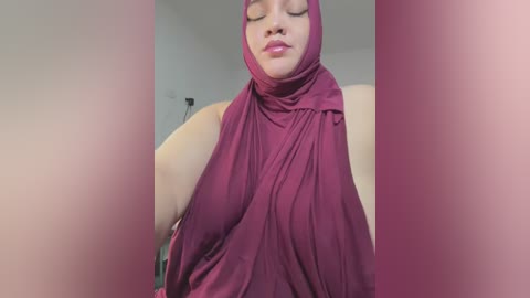 Video of a light-skinned woman with a medium build wearing a deep maroon hijab and matching clothing, eyes closed in a serene expression. Background is blurred with a soft pink hue.