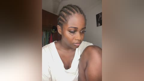 Media: Video of a young Black woman with braided hair, wearing a white top, gazing downward in a dimly lit room with hanging clothes and a framed photo on the wall.