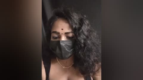 Media: Video of a South Asian woman with medium brown skin, wearing a black face mask, black saree, and a gold necklace, with curly black hair partially covering her forehead. Dark background.