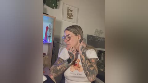 Media: Video of a tattooed woman with glasses, wearing a white t-shirt featuring a cartoon dog, sitting in a dimly lit room with a shelf displaying sex toys and framed art.