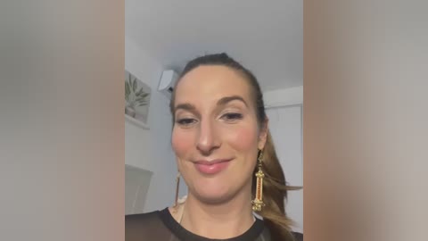 Media: Video of a smiling woman with fair skin and brown hair tied back, wearing gold earrings and a black top, indoors with white walls and a leafy plant.