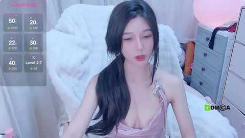 Media: A video of an Asian woman with long black hair, fair skin, and red lipstick, wearing a revealing pink lace camisole, sitting on a white bed in a dimly lit room.