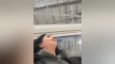 Media: Video of a woman with fair skin and dark hair in a green hoodie, standing under a modern, industrial ceiling with metal beams and glass panels, looking upward.