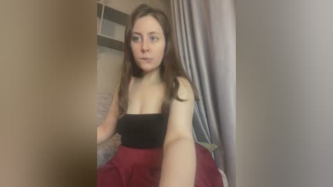 Media: Video of a young woman with light skin, long brown hair, wearing a black strapless top and red skirt, sitting indoors against a beige wall with a bookshelf and grey curtains in the background.