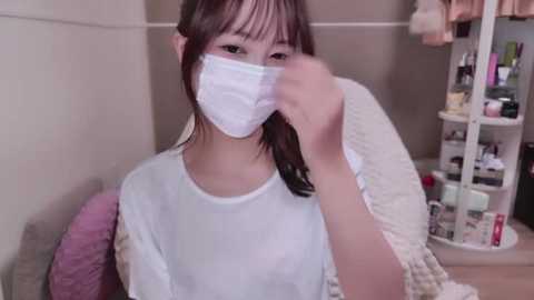 Media: Video of an Asian woman with light skin and straight brown hair, wearing a white mask, white T-shirt, and beige sweater, seated indoors. Background shows a cluttered makeup vanity with various cosmetics.