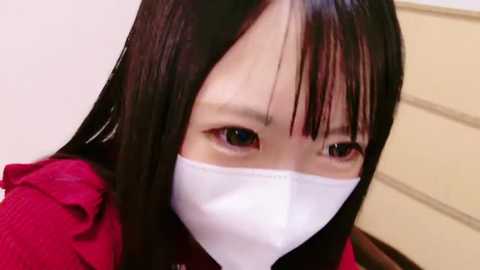 Video of an Asian woman with long black hair, wearing a white surgical mask and a red jacket, standing indoors with beige walls and a wooden dresser in the background.