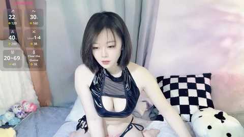 Media: A video of an East Asian woman with shoulder-length black hair, wearing a shiny black latex bikini, sitting on a bed with checkered pillows, in a softly lit room.