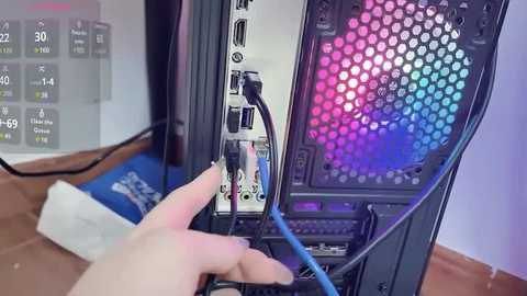 Video of a person plugging a blue cable into a computer tower with a hexagonal, multicolored LED screen. Background includes a whiteboard with temperature readings and a blue bag.