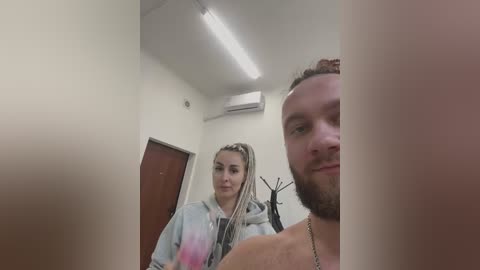 Media: Video of a woman with blonde dreadlocks in a tie-dye hoodie and a man with a beard, smiling, in a small, dimly lit room with white walls, a brown door, and an air conditioning unit.