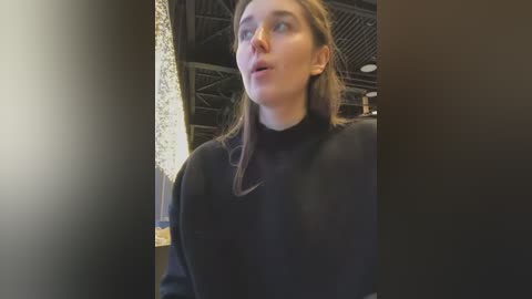 Video of a young Caucasian woman with fair skin, straight brown hair, and a round face, wearing a black turtleneck sweater. She has a serious expression, standing in an industrial setting with exposed beams and metallic textures in the background.