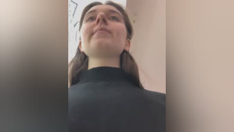 A low-angle video of a young woman with fair skin, brown hair, and glasses, wearing a black turtleneck, standing in a narrow corridor with beige walls.