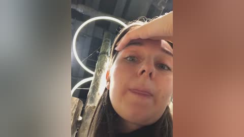 Video of a young woman with fair skin, green eyes, and shoulder-length brown hair, wearing a brown jacket, touching her forehead. Background shows a circular, glowing light fixture and industrial pipes.