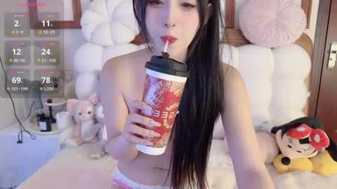 Media: Video of a young Asian woman with long black hair, wearing white panties, holding a red Starbucks cup, in a pink and white themed bedroom with plush toys and a bed.